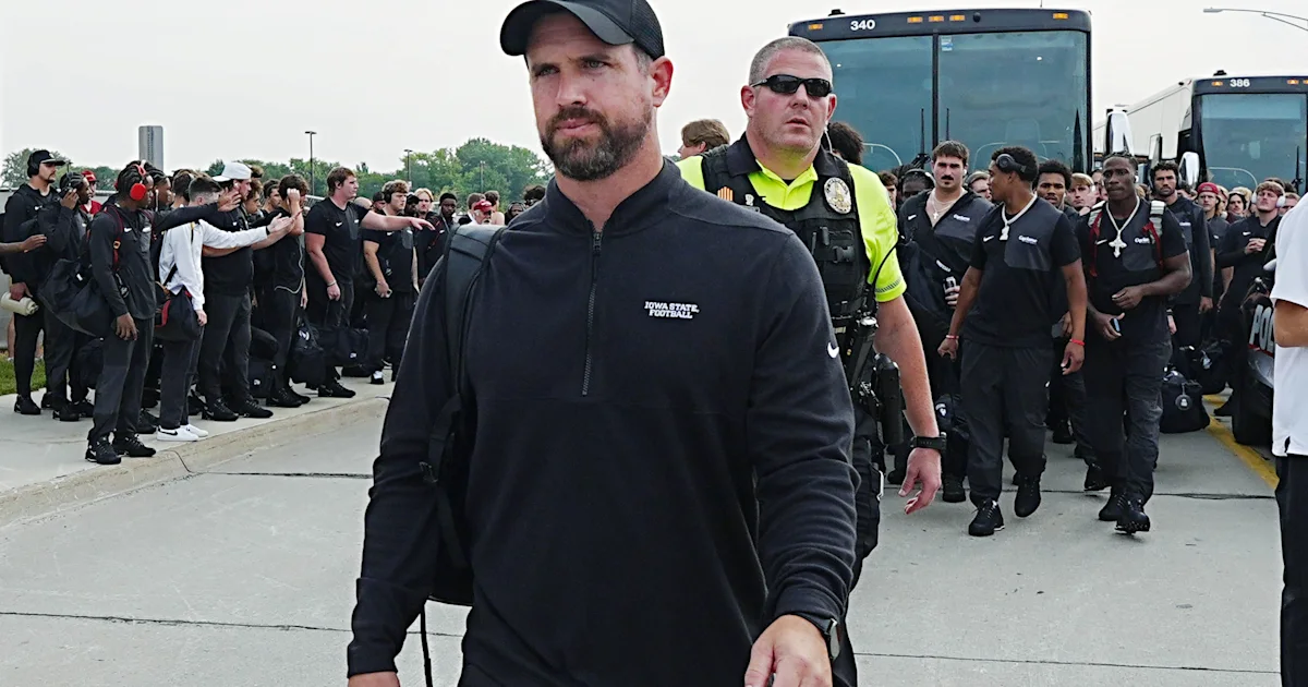 A man in an Iowa State football pullover walks past a security officer and a crowd beside buses.