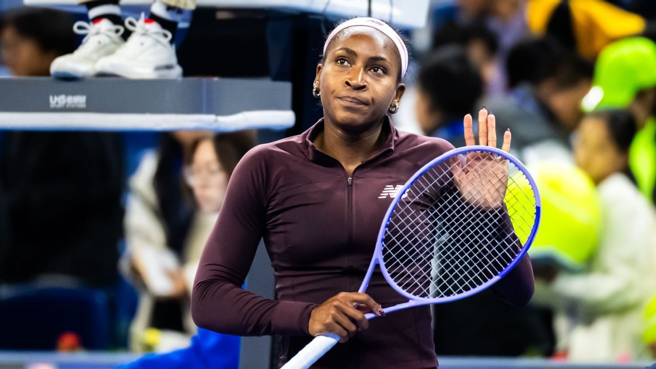Tennis player holding a racket raises her hand amid a blurred crowd.