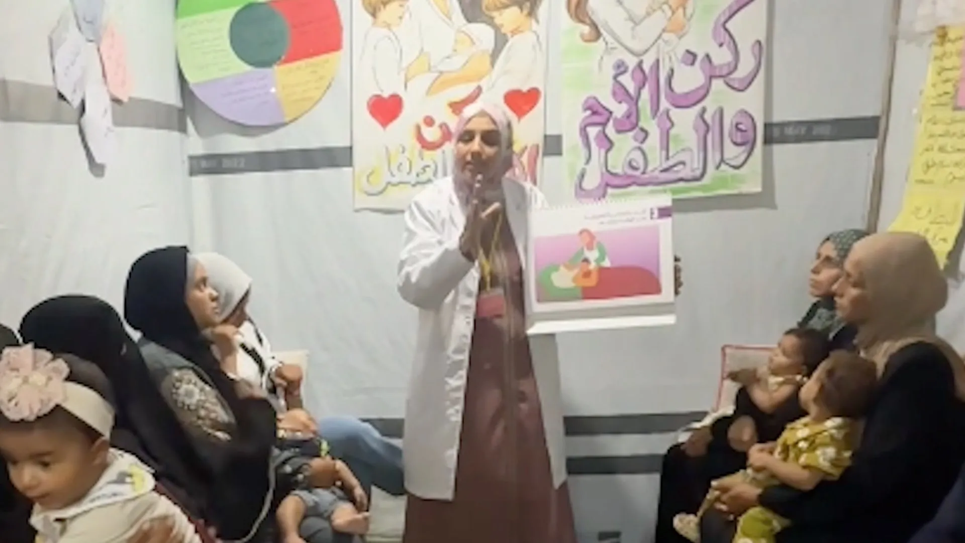 A woman in a white coat speaks to seated mothers holding babies, with illustrated posters behind her.
