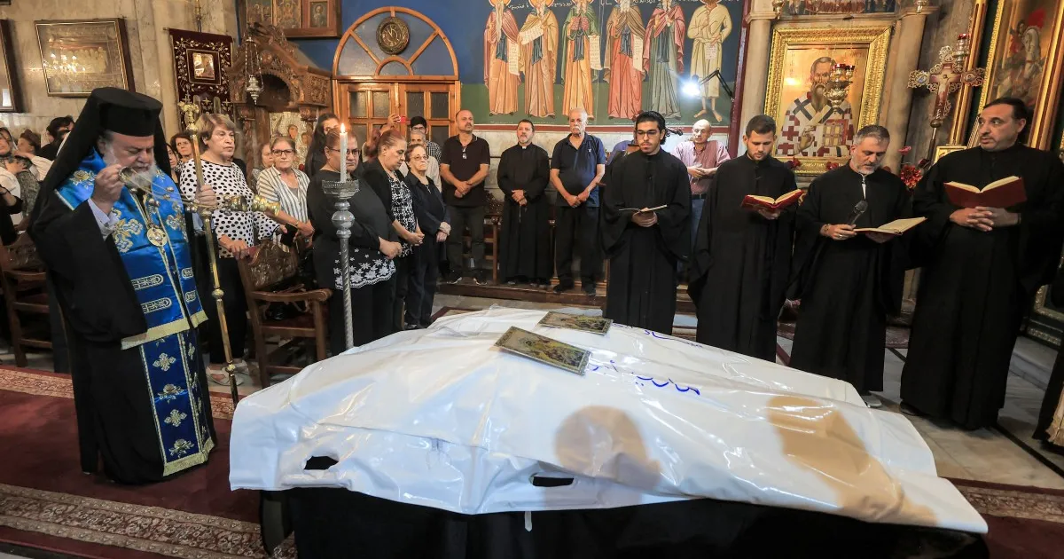 Clergy and mourners gather around a white-covered body during a service inside an ornate church.