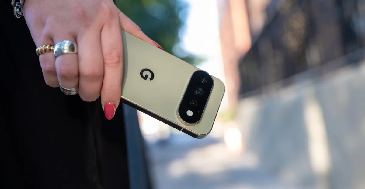 A hand with rings and red nail polish holds a pale-colored smartphone with a vertical rear camera bar.