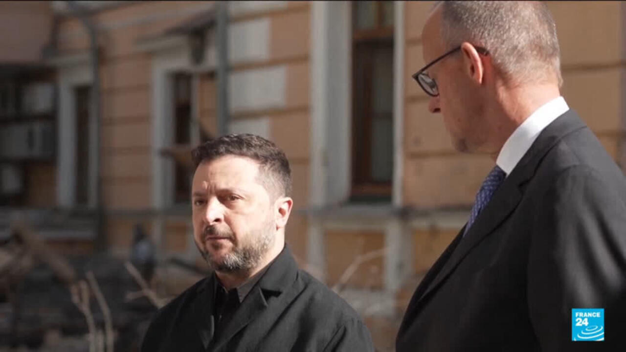 Two men in dark suits stand outside a damaged building, with debris visible behind them.