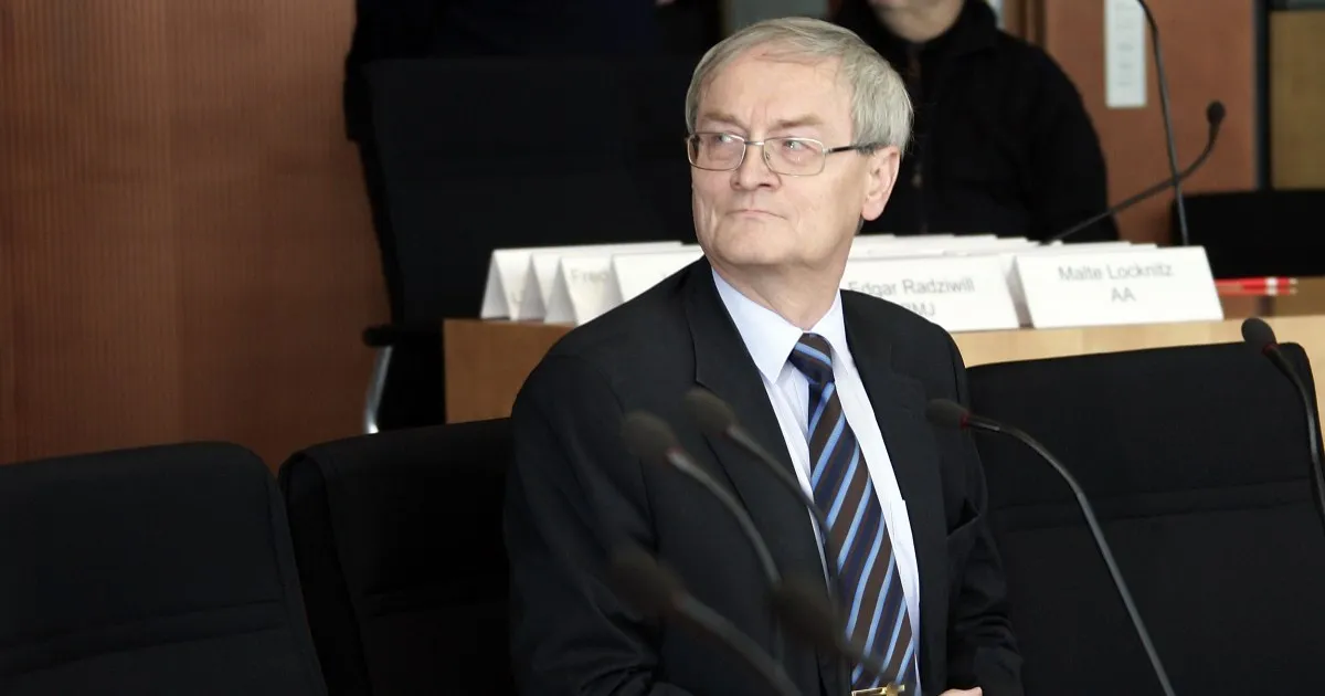 Older man in a dark suit and striped tie sits at a conference table among microphones.