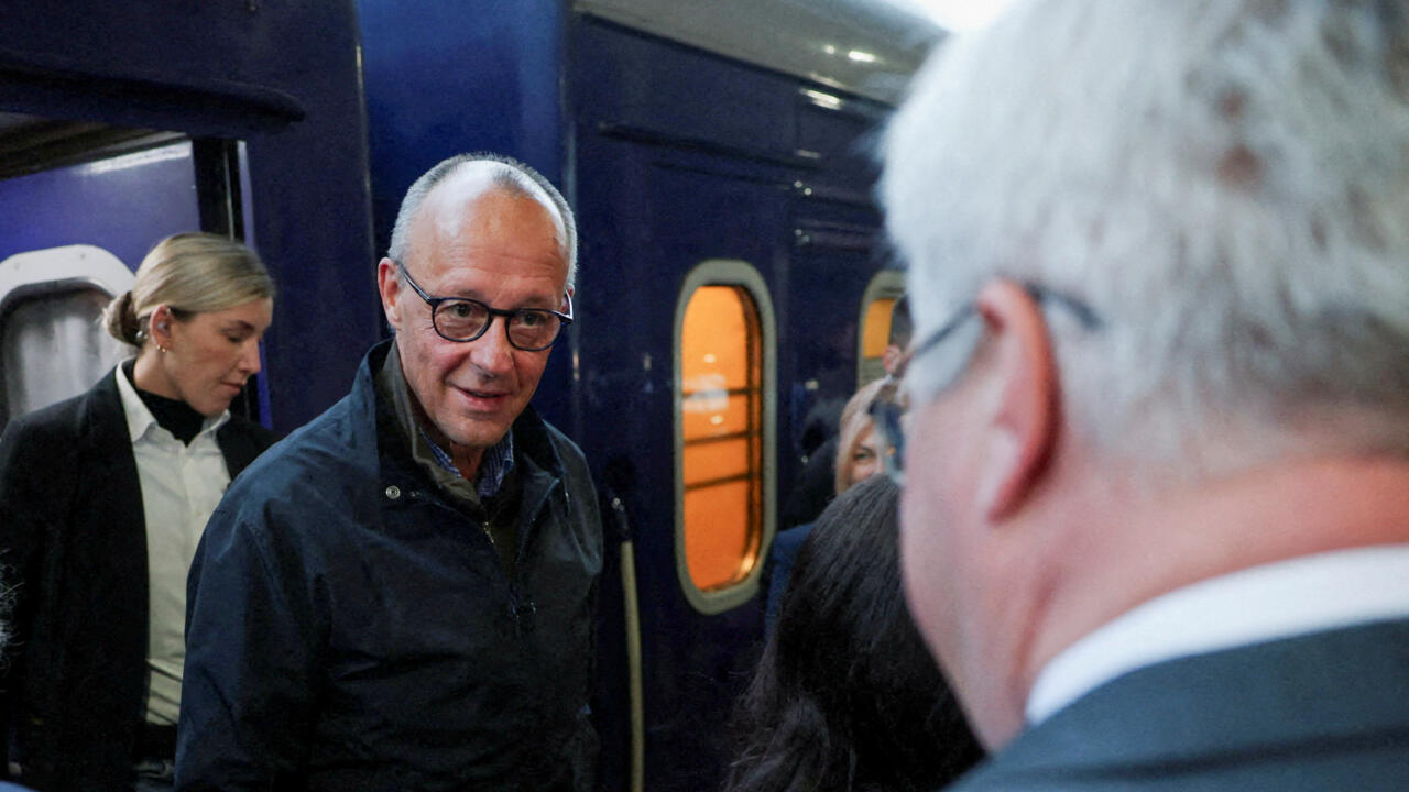 A bespectacled man in a dark jacket speaks with a white-haired man aboard a train.