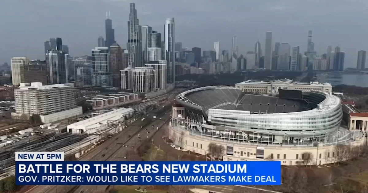 Aerial view of an oval stadium beside rail yards and a dense city skyline under an overcast sky.