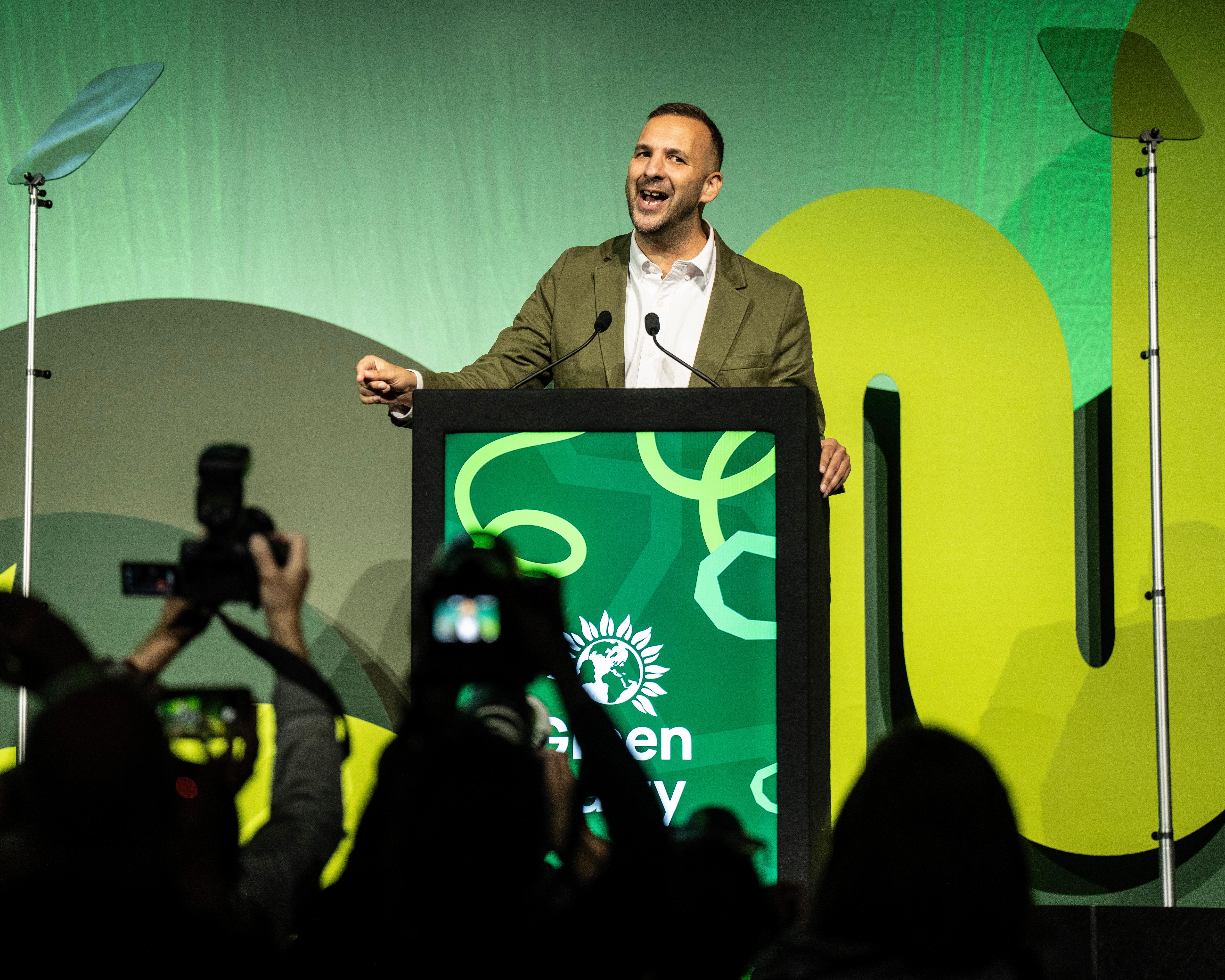 A man speaks at a green lectern as photographers in the foreground take pictures.