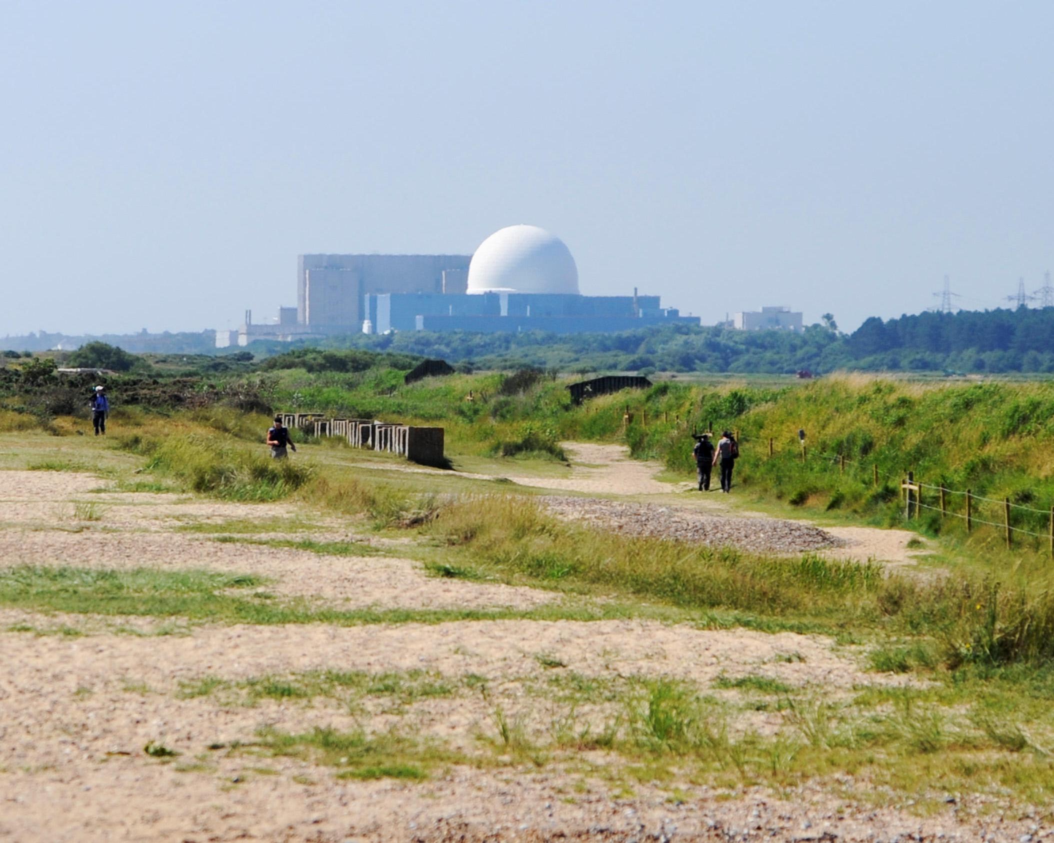 People walk along a sandy path through grassland, with a large industrial building and white dome in the distance.