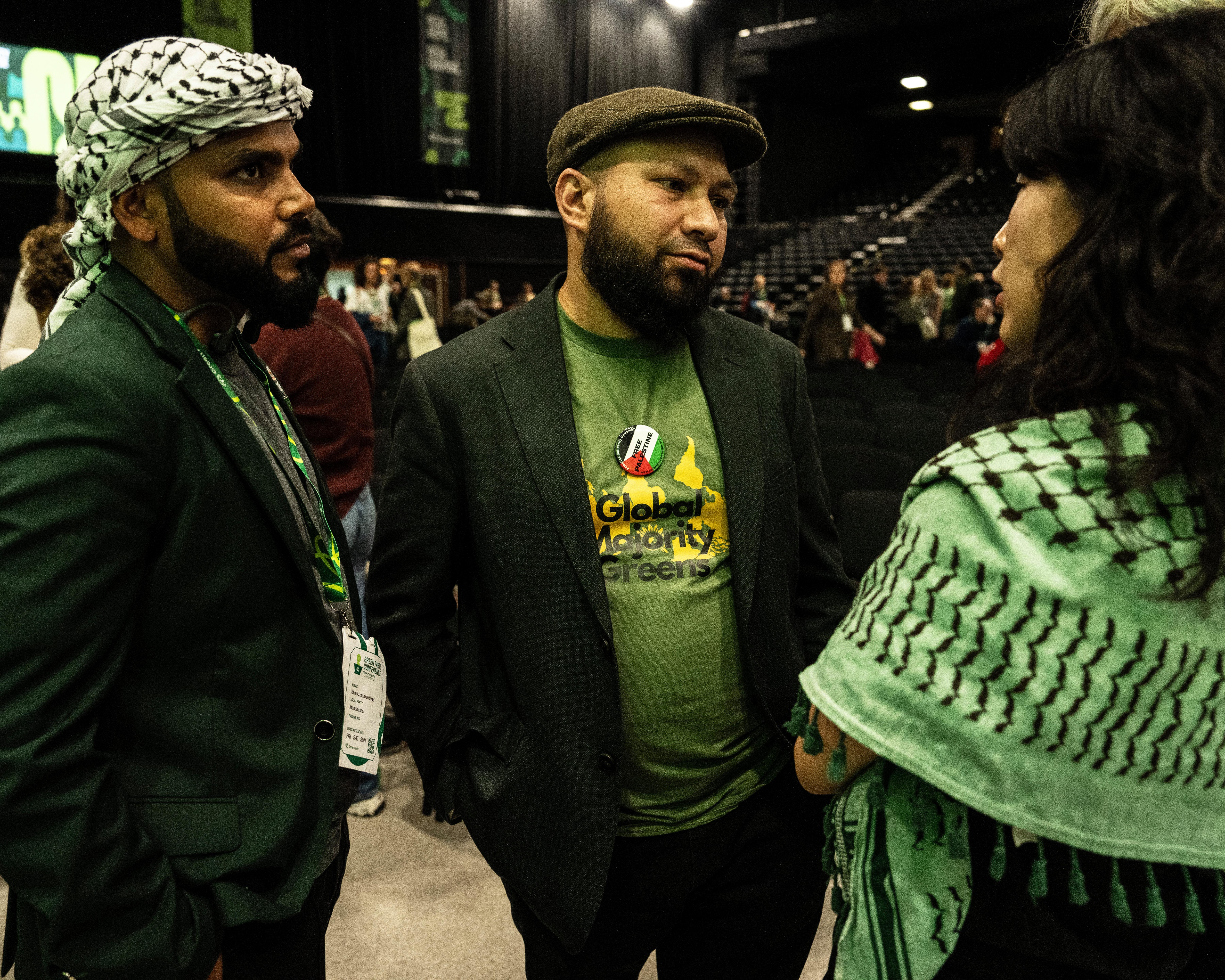 Three people converse at a conference; two wear keffiyehs and the central man wears a Green party T-shirt.