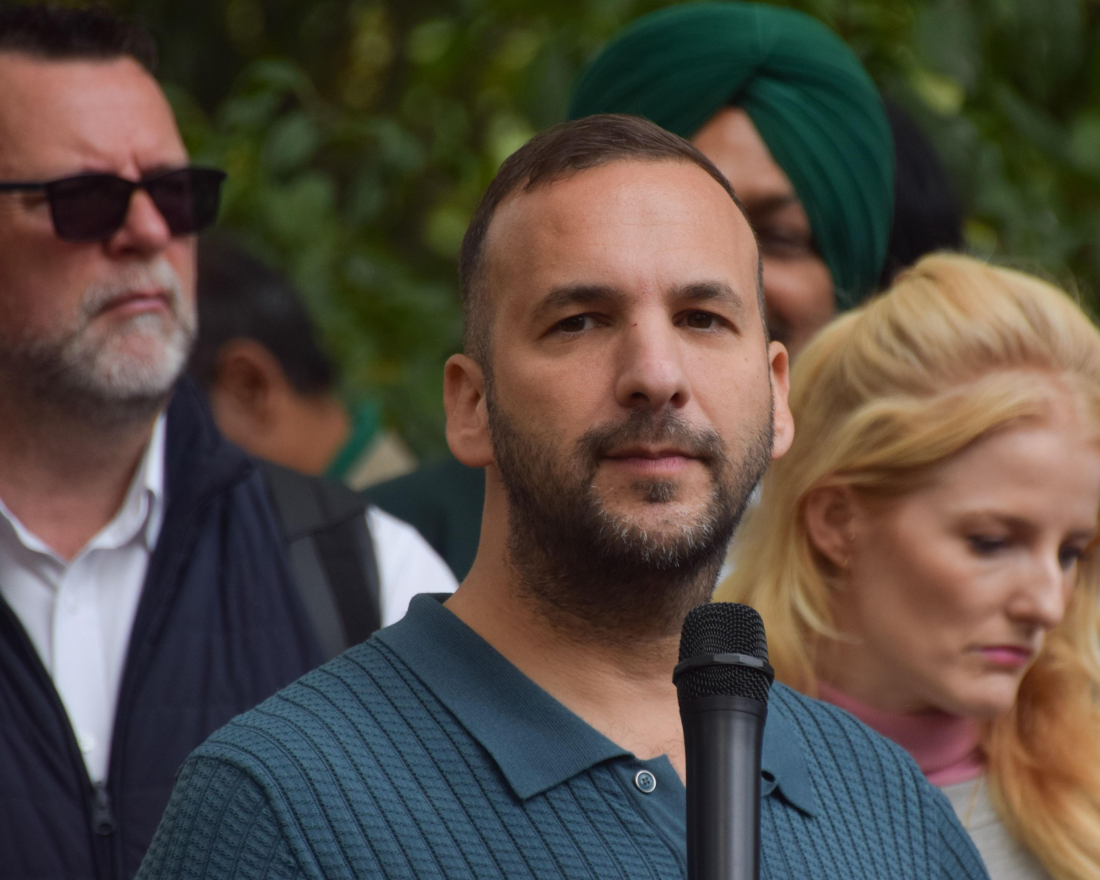 A bearded man holds a microphone among several people, including a man in sunglasses and a woman.