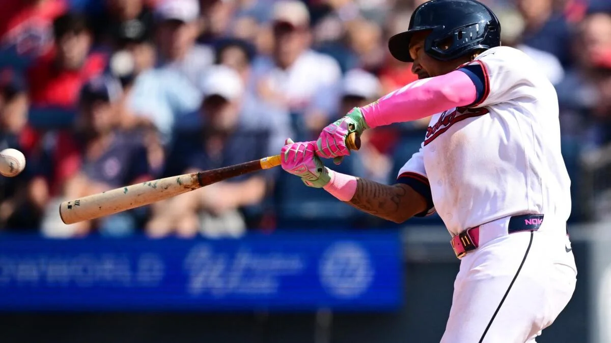 A baseball player in a white uniform and pink gloves swings at a pitch as the ball approaches.