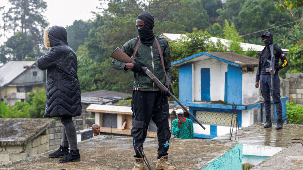Masked armed men stand on a rooftop near a woman and a small blue-and-white building.
