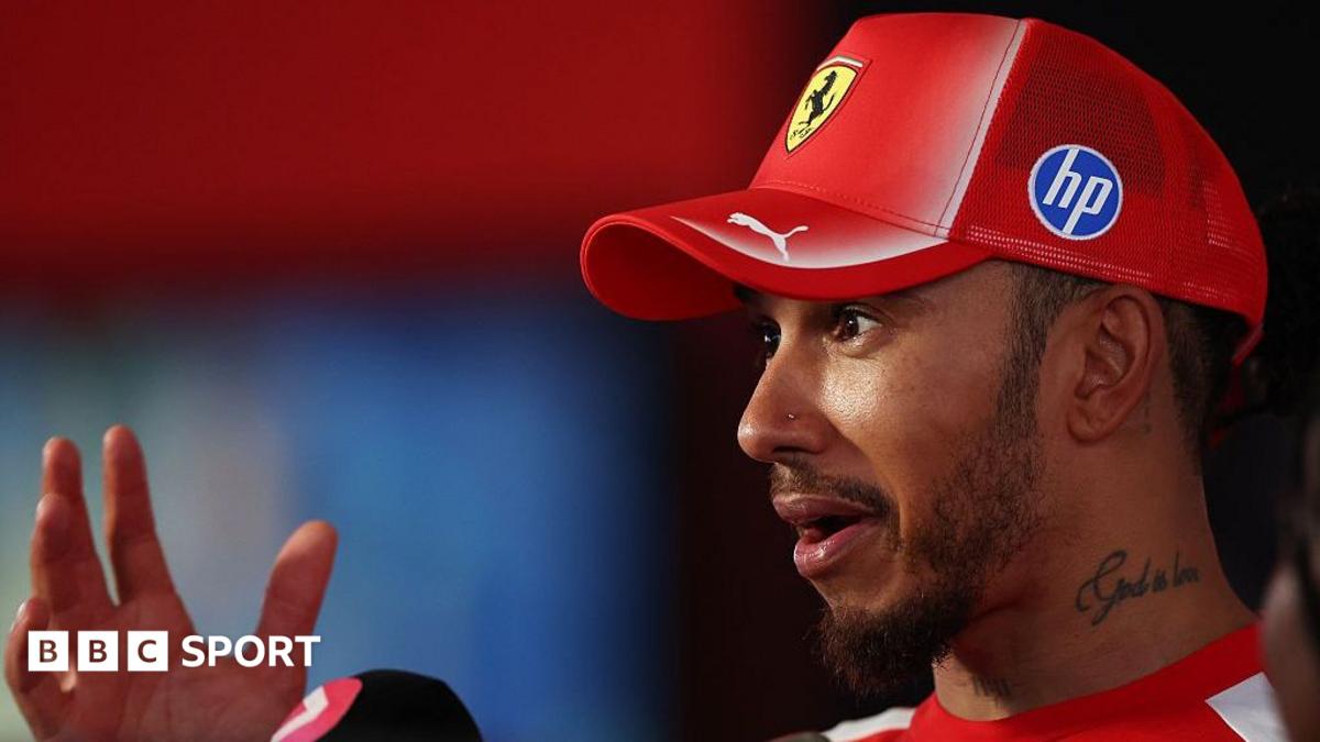 Lewis Hamilton wears a red Ferrari cap and raises one hand during an interview.