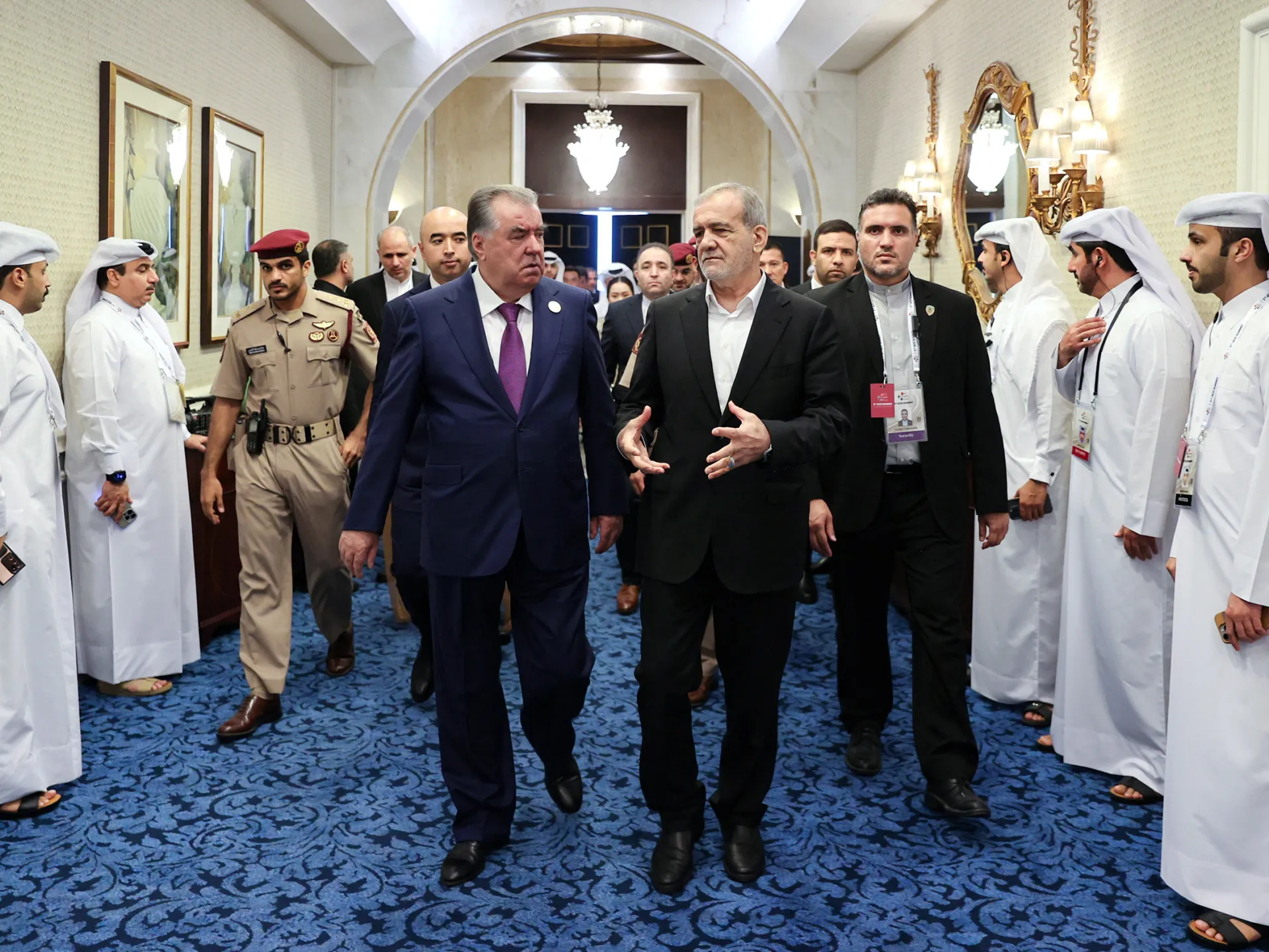 Two suited officials walk through an ornate corridor as uniformed personnel and attendants stand on either side.