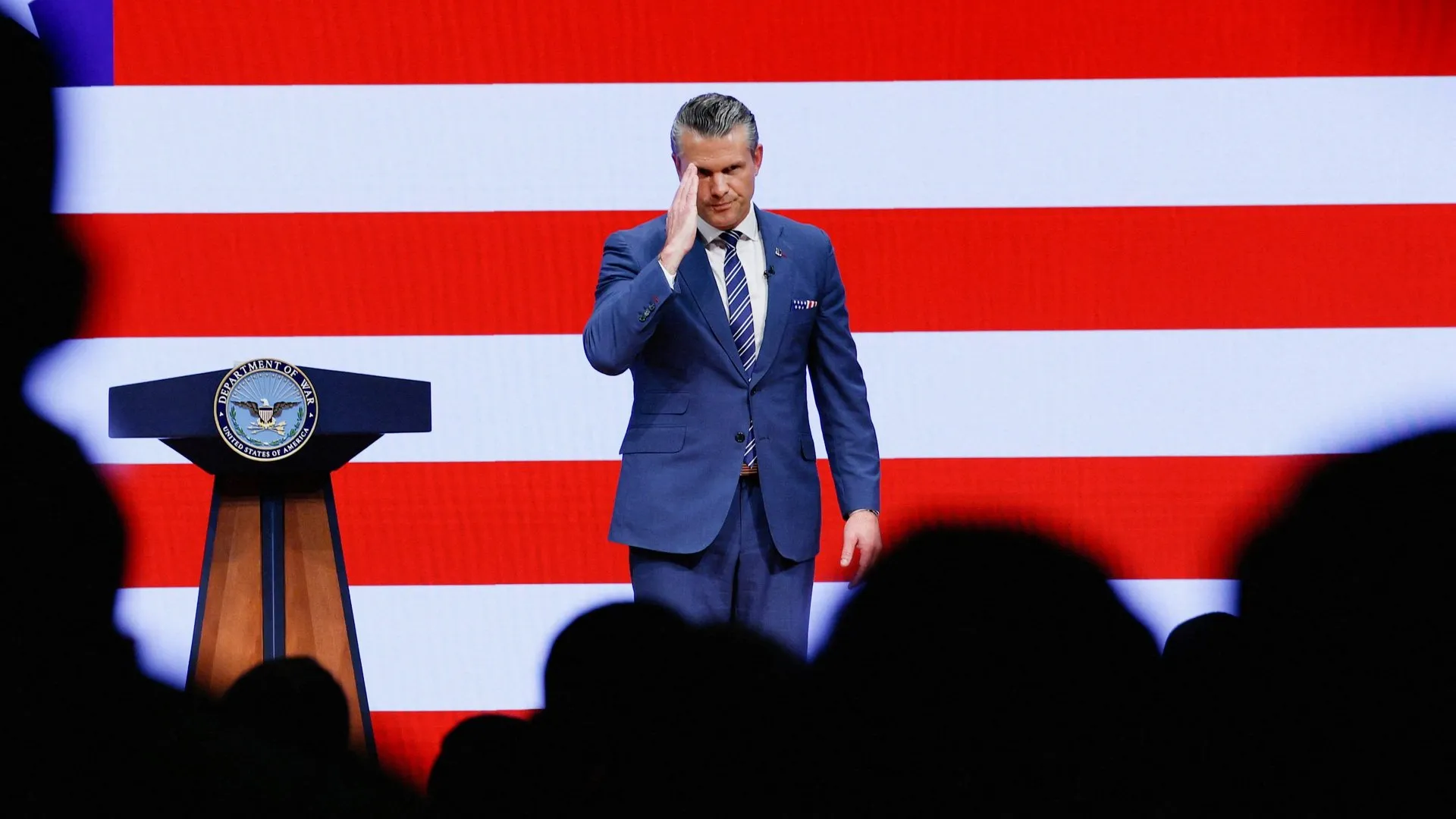 A man in a blue suit raises his hand beside a Defense Department podium, with an American flag behind him.