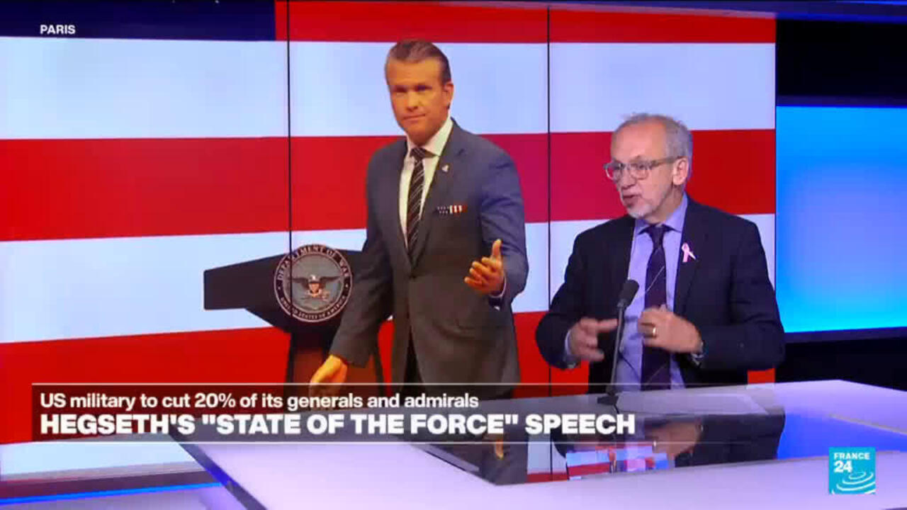 Two suited men stand beside a podium in a studio with red, white, and blue screens.