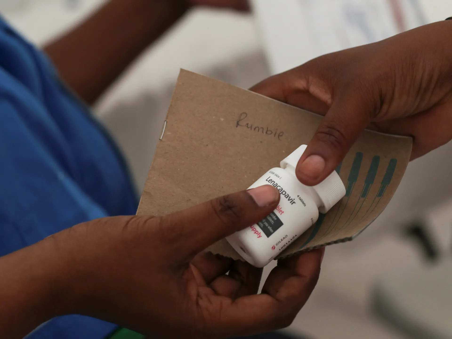Hands hold a bottle of lenacapavir beside a cardboard booklet labeled “Rumble.”