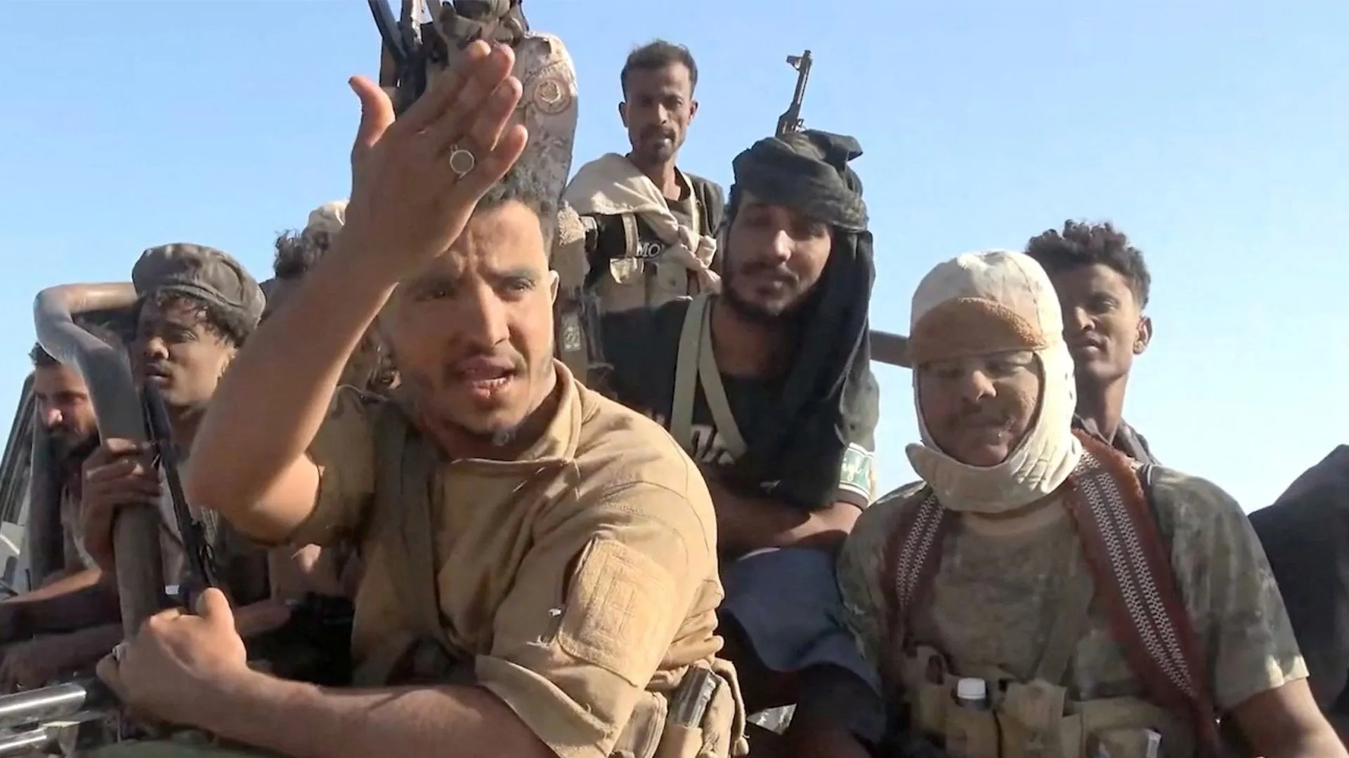 Armed men ride in a vehicle under a clear sky, one raising his hand toward the camera.