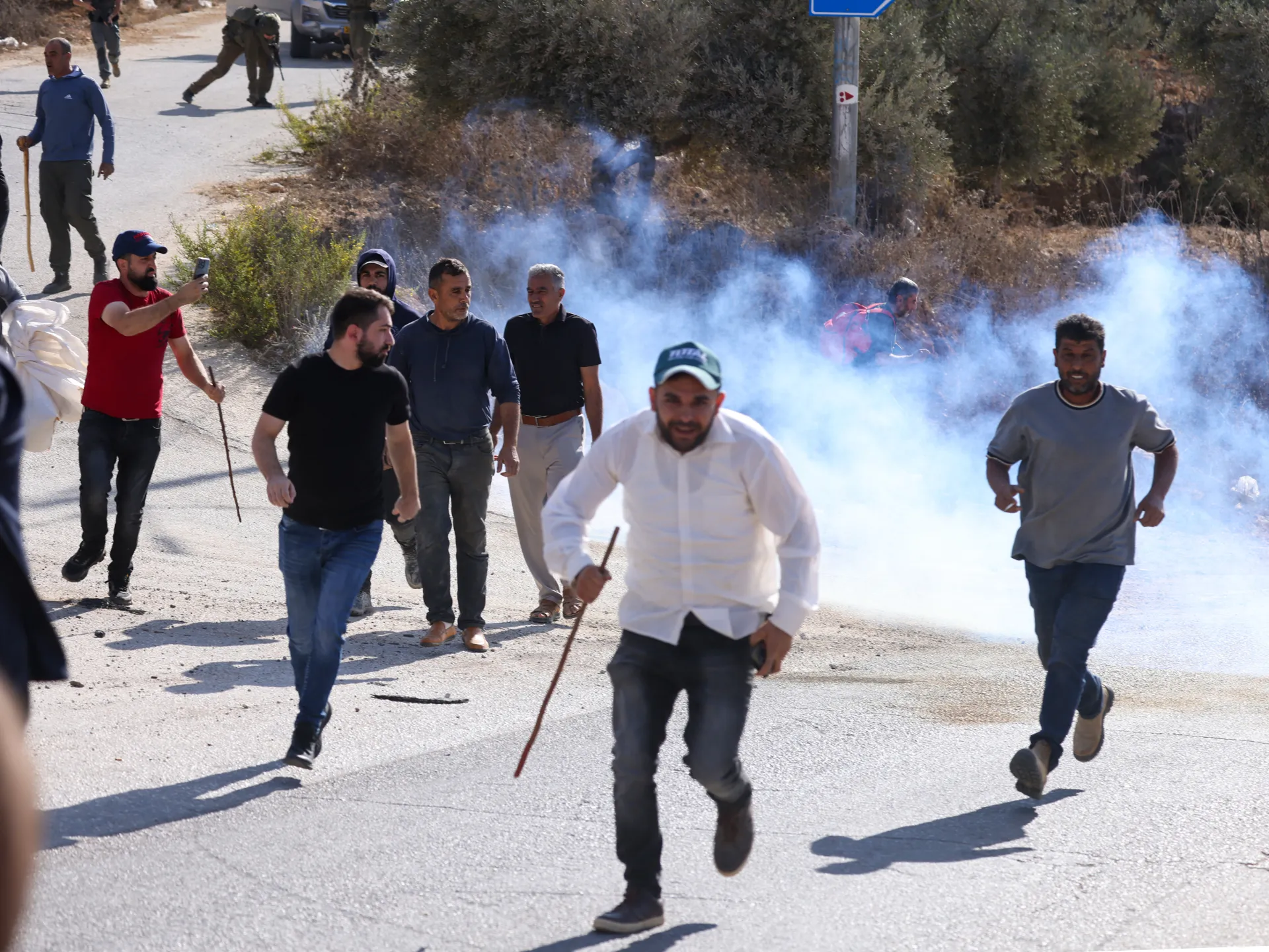 Several men run along a road as smoke billows nearby, with armed personnel visible in the background.