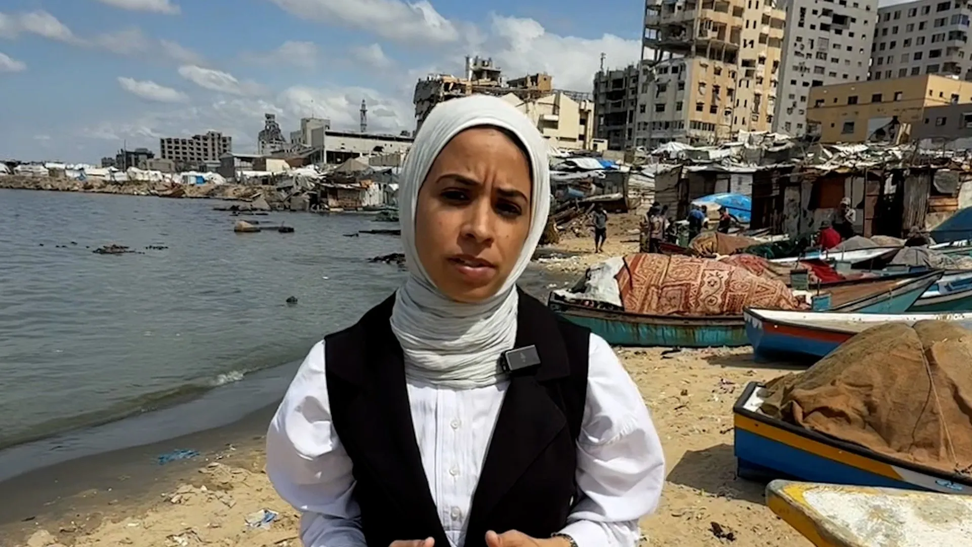 A woman in a white hijab stands beside boats, damaged buildings, and debris along a shoreline.