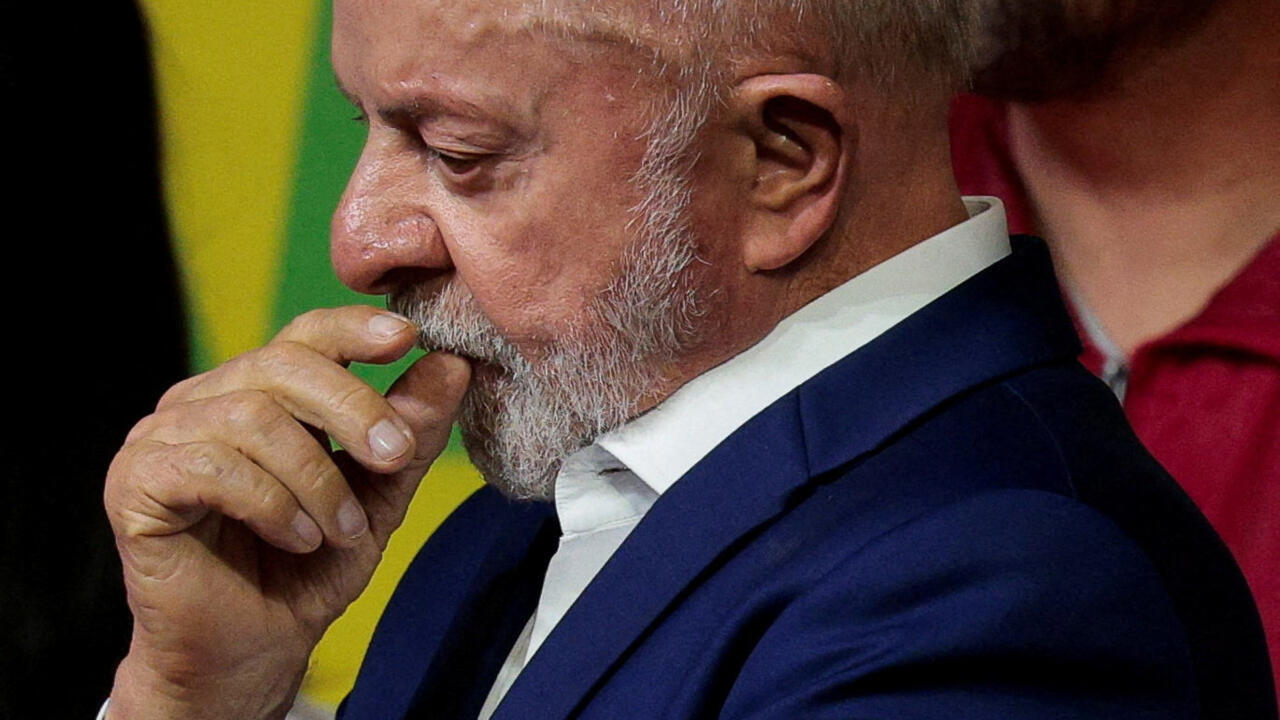 A gray-bearded man in a dark suit rests his fingers against his lips, looking downward.