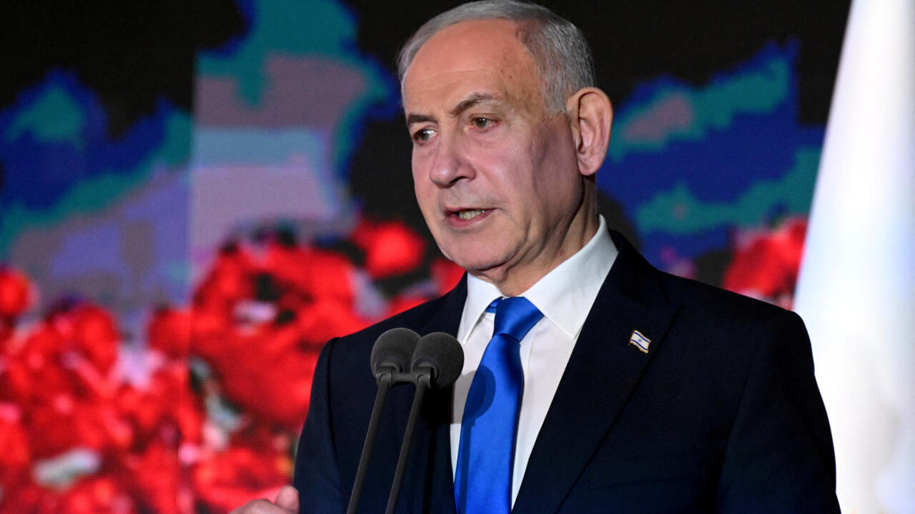 A gray-haired man in a dark suit and blue tie speaks at a pair of microphones.