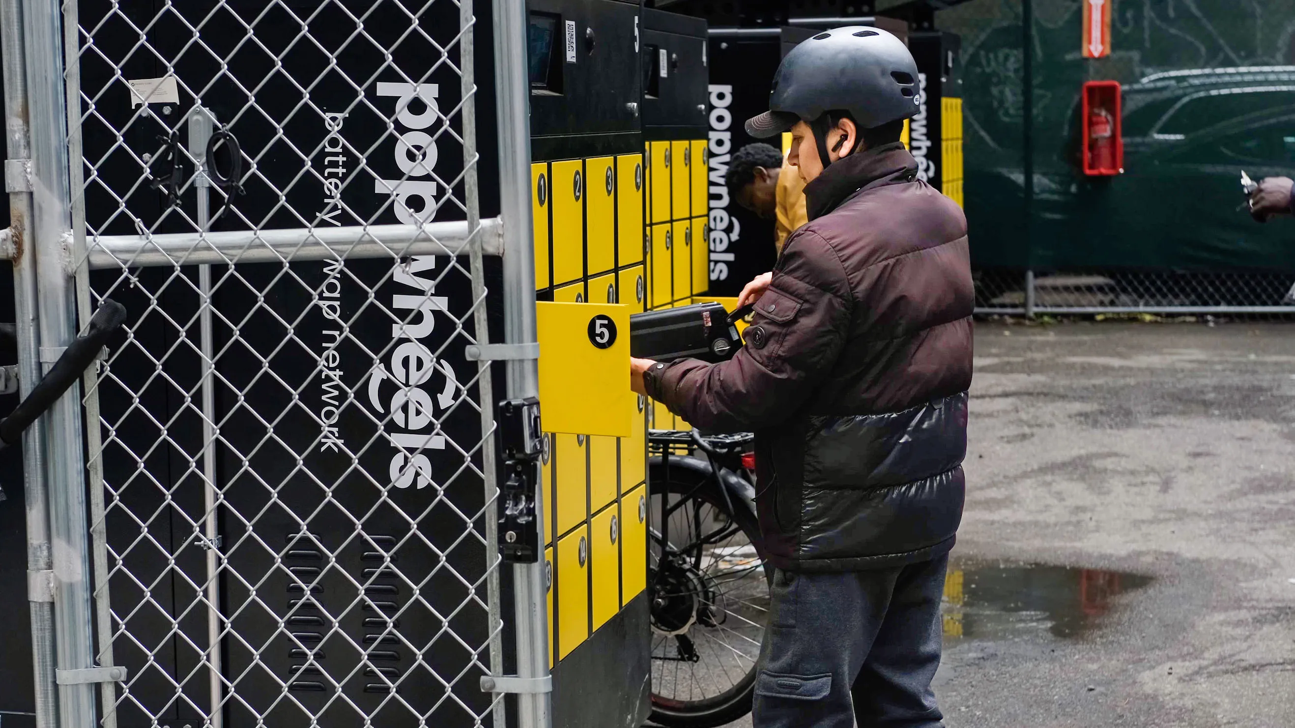 A helmeted person uses a battery locker beside a bicycle behind a chain-link fence.