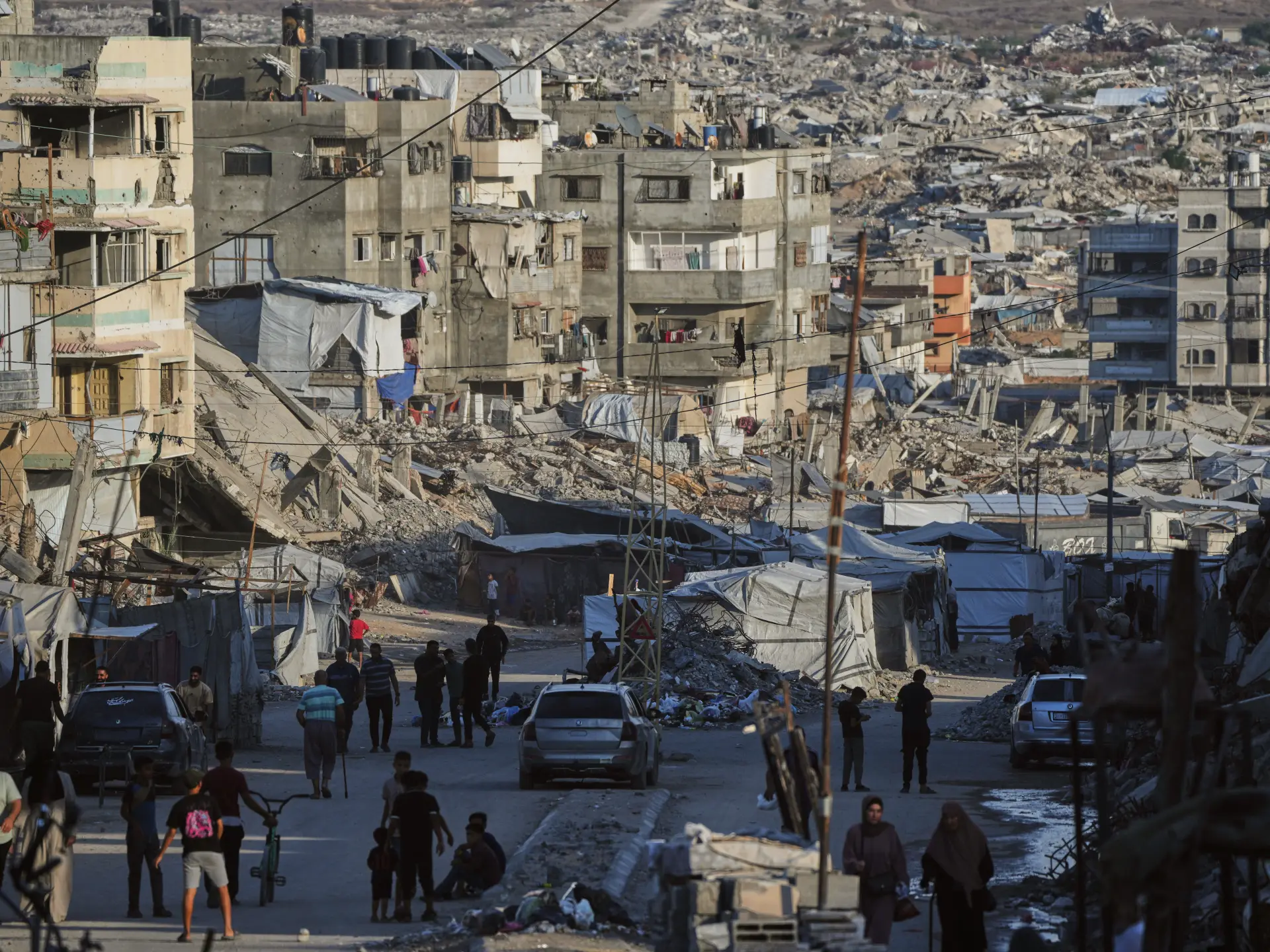 People walk past tents and heavily damaged buildings amid extensive rubble and debris.