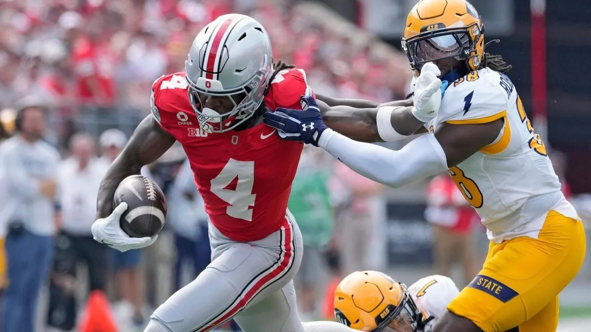 Ohio State player in red carries the football as a player in white and yellow reaches toward him.