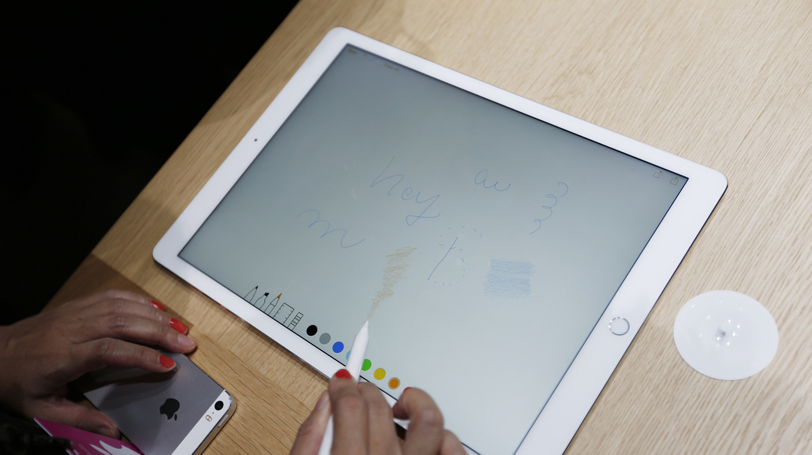 A person uses an Apple Pencil on an iPad screen beside an iPhone on a wooden table.