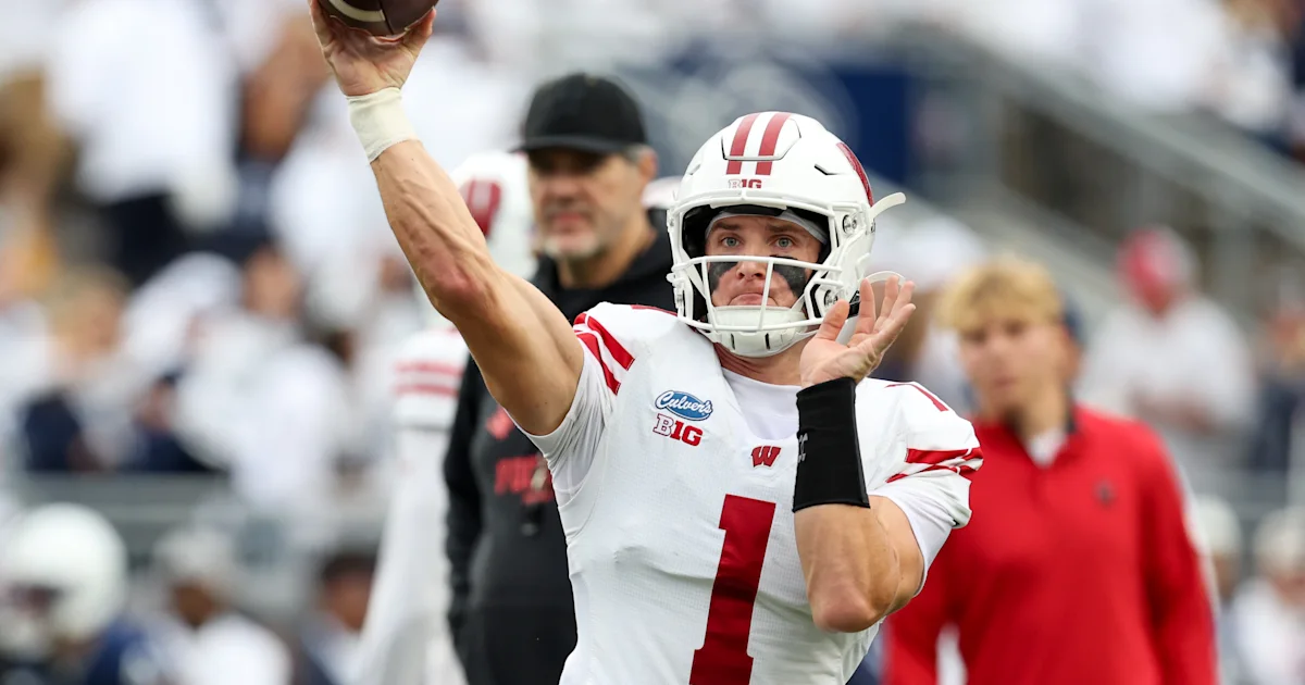 Wisconsin quarterback in a white and red uniform raises a football to throw amid blurred sideline figures.