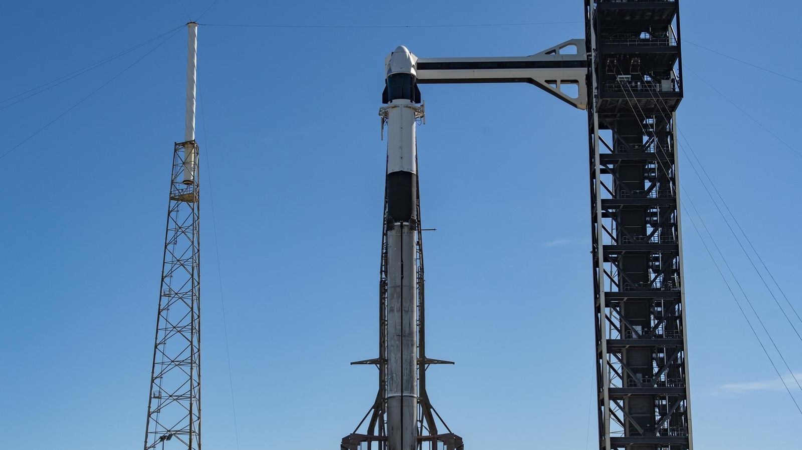 A crew capsule atop a tall rocket stands beside a launch tower under a clear blue sky.