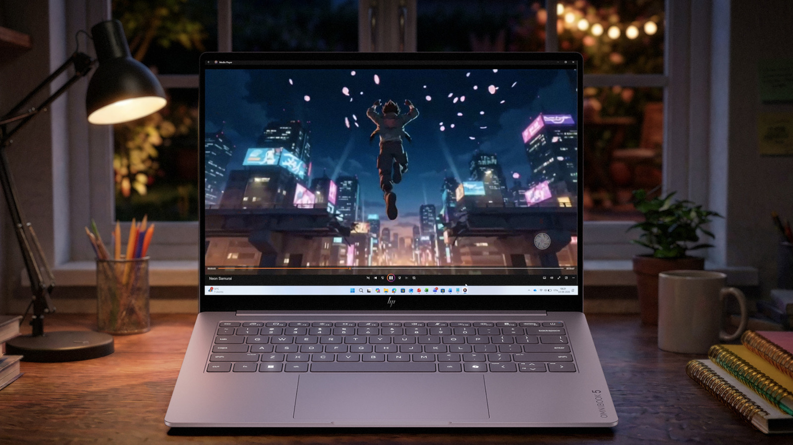 HP OmniBook laptop playing an animated video on a desk beside a lamp and stationery