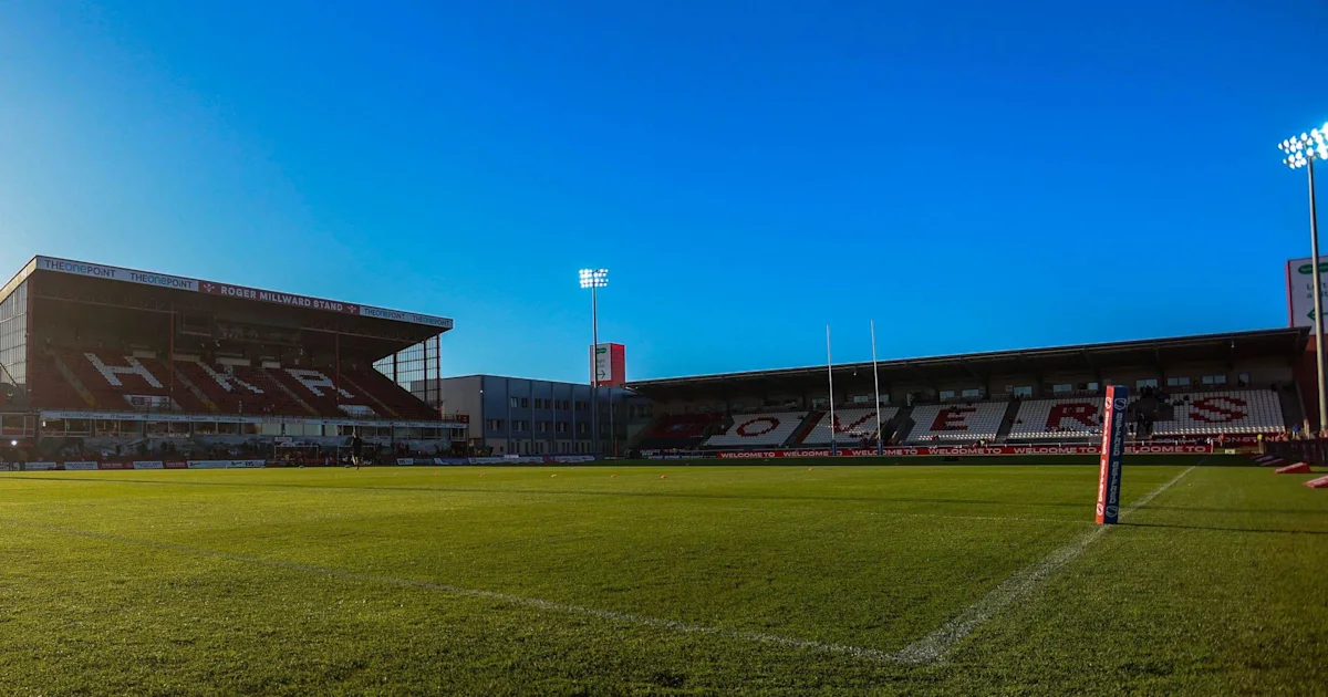 Empty rugby league pitch with goalposts, stadium stands and floodlights beneath a clear blue sky.