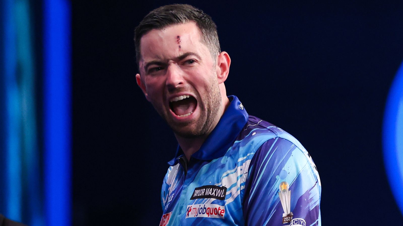 A darts player in a blue patterned shirt shouts, with a small mark visible on his forehead.