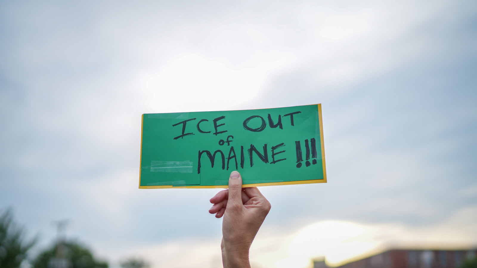 A hand holds a green sign reading “ICE out of Maine!!!” against a cloudy sky.