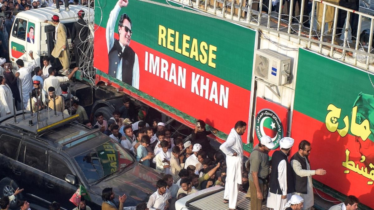 Crowd surrounds vehicles beside a large green and red banner reading “Release Imran Khan.”