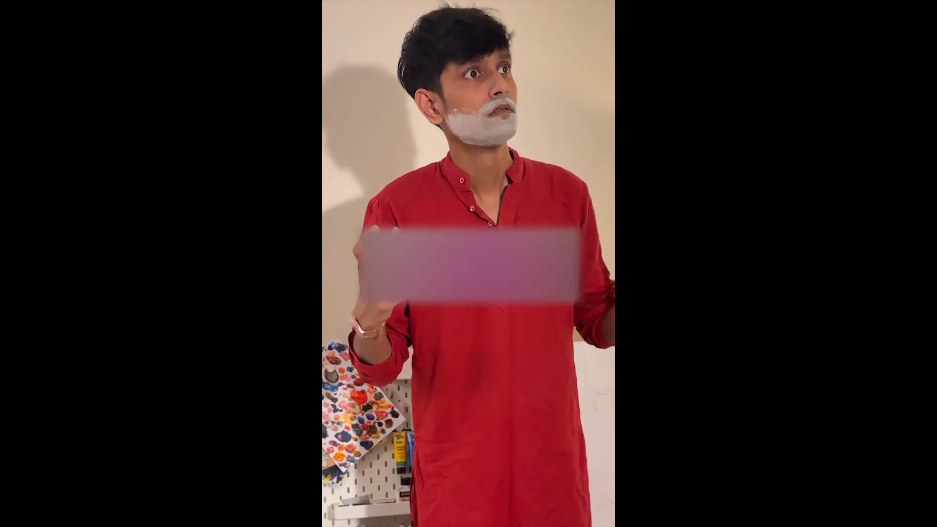 Man in a red kurta with white cream on his face gestures while looking upward.