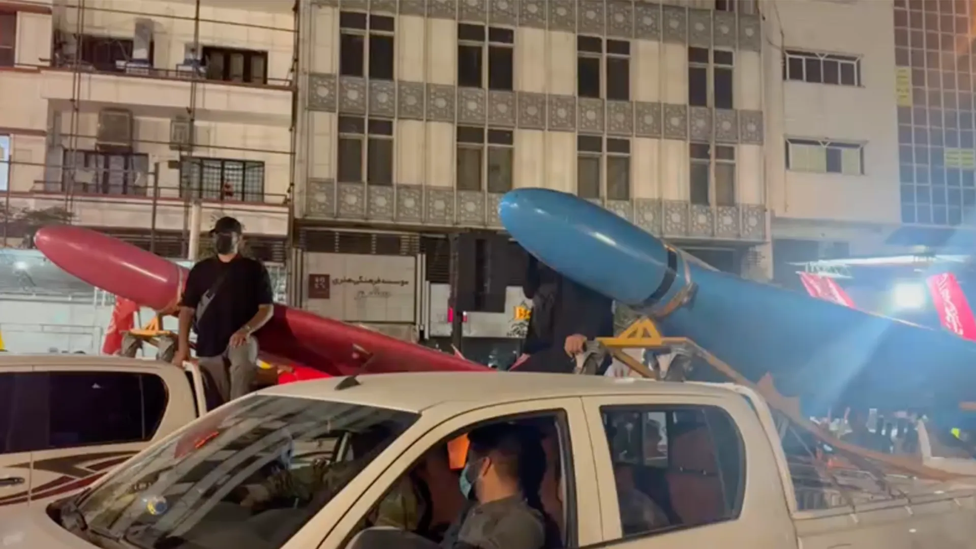 Blue and red drone-shaped displays are carried past cars and onlookers on a city street at night.
