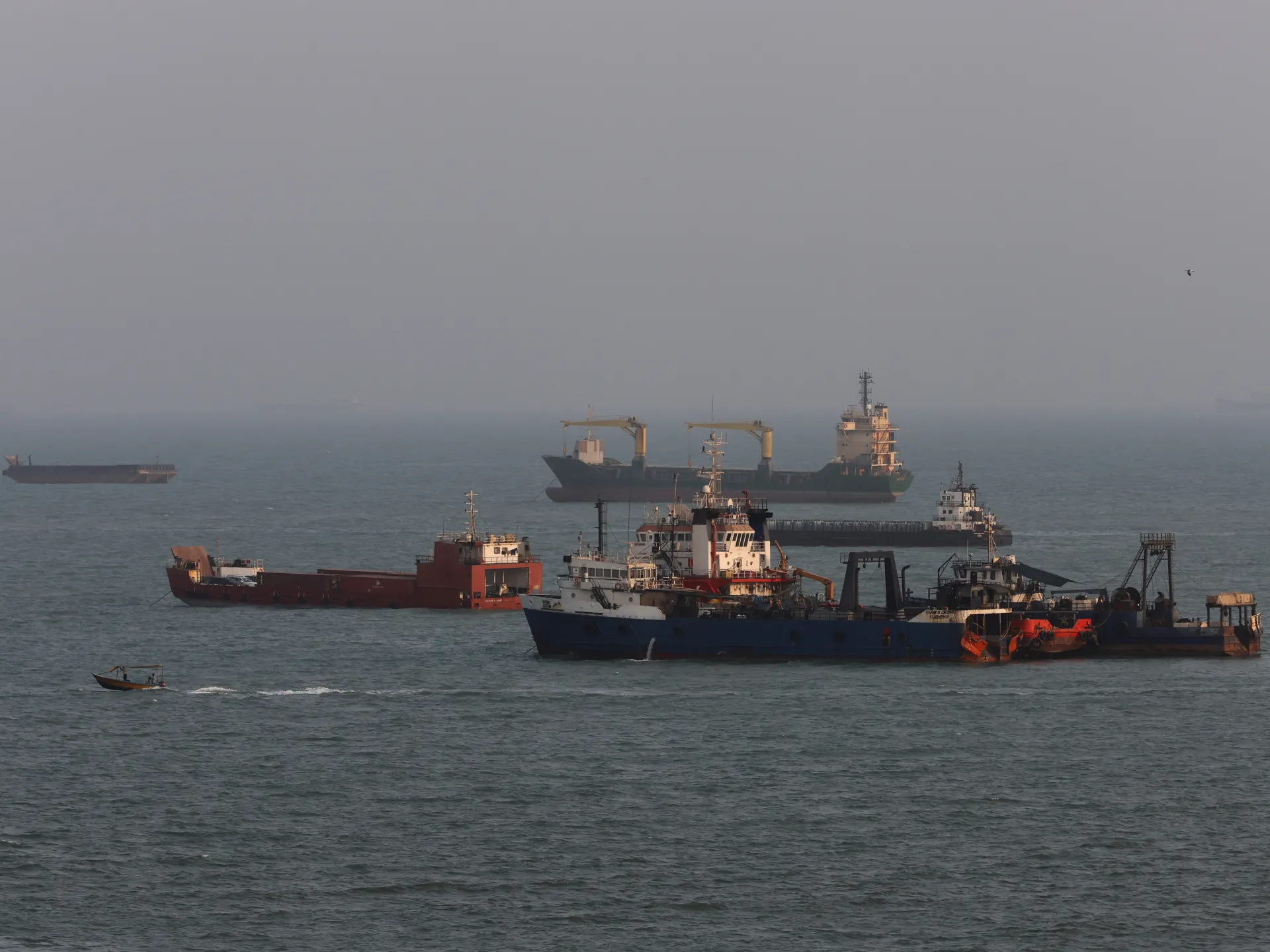 Several cargo and work vessels sit on hazy water, with a small boat passing in the foreground.