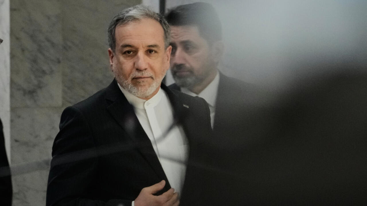 A gray-haired man in a dark suit walks indoors, with another man visible behind him through glass.