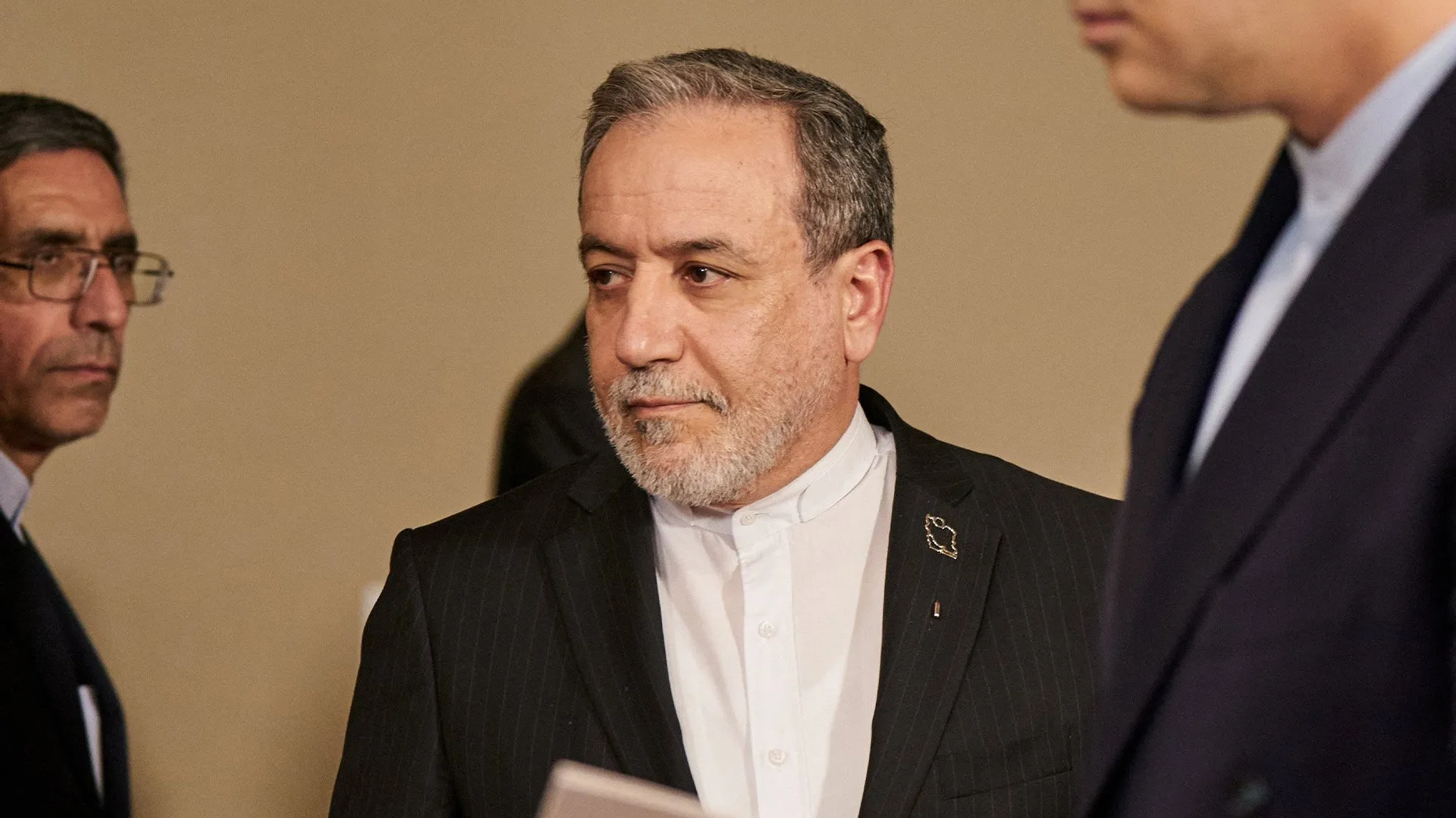 A gray-haired man in a dark suit stands between two other men indoors.