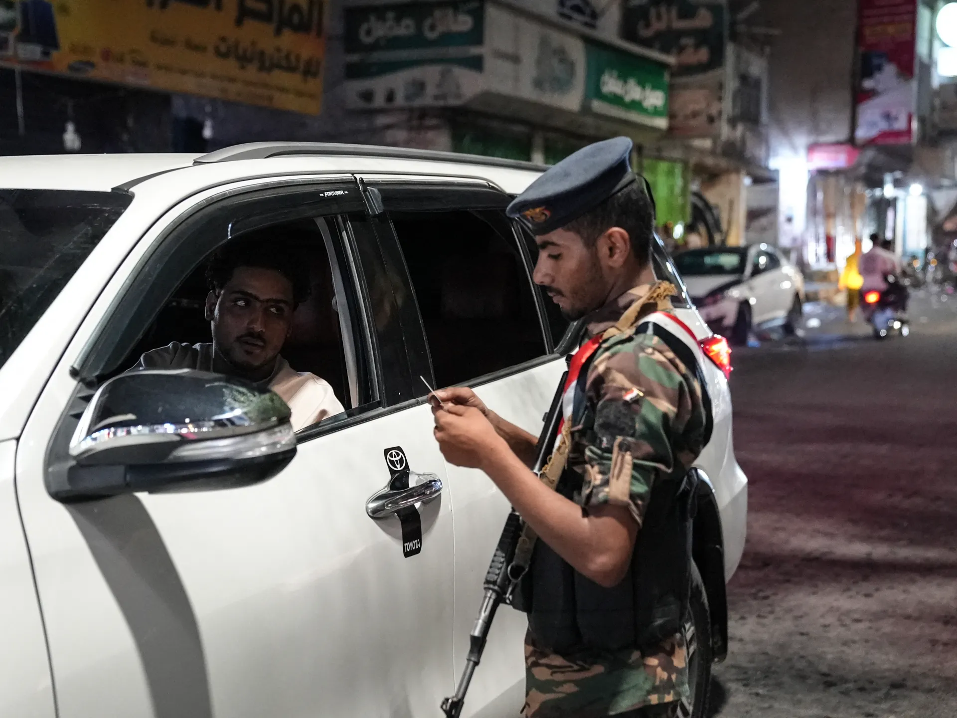 A uniformed armed man checks a document beside a driver in a white SUV at night.