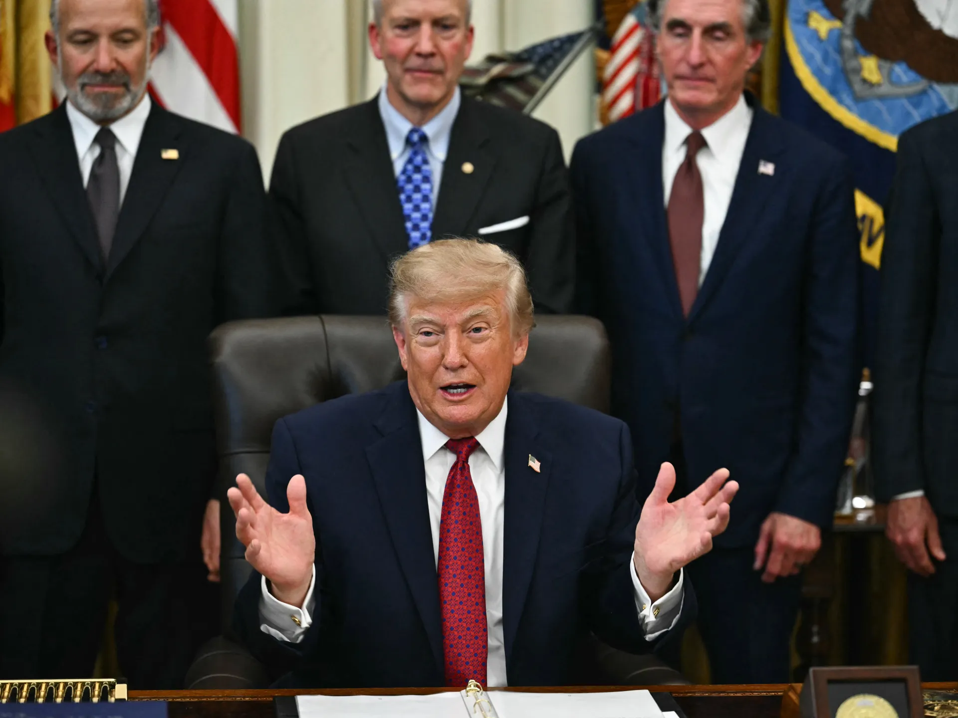 Donald Trump speaks with both hands raised as several suited men stand behind him.