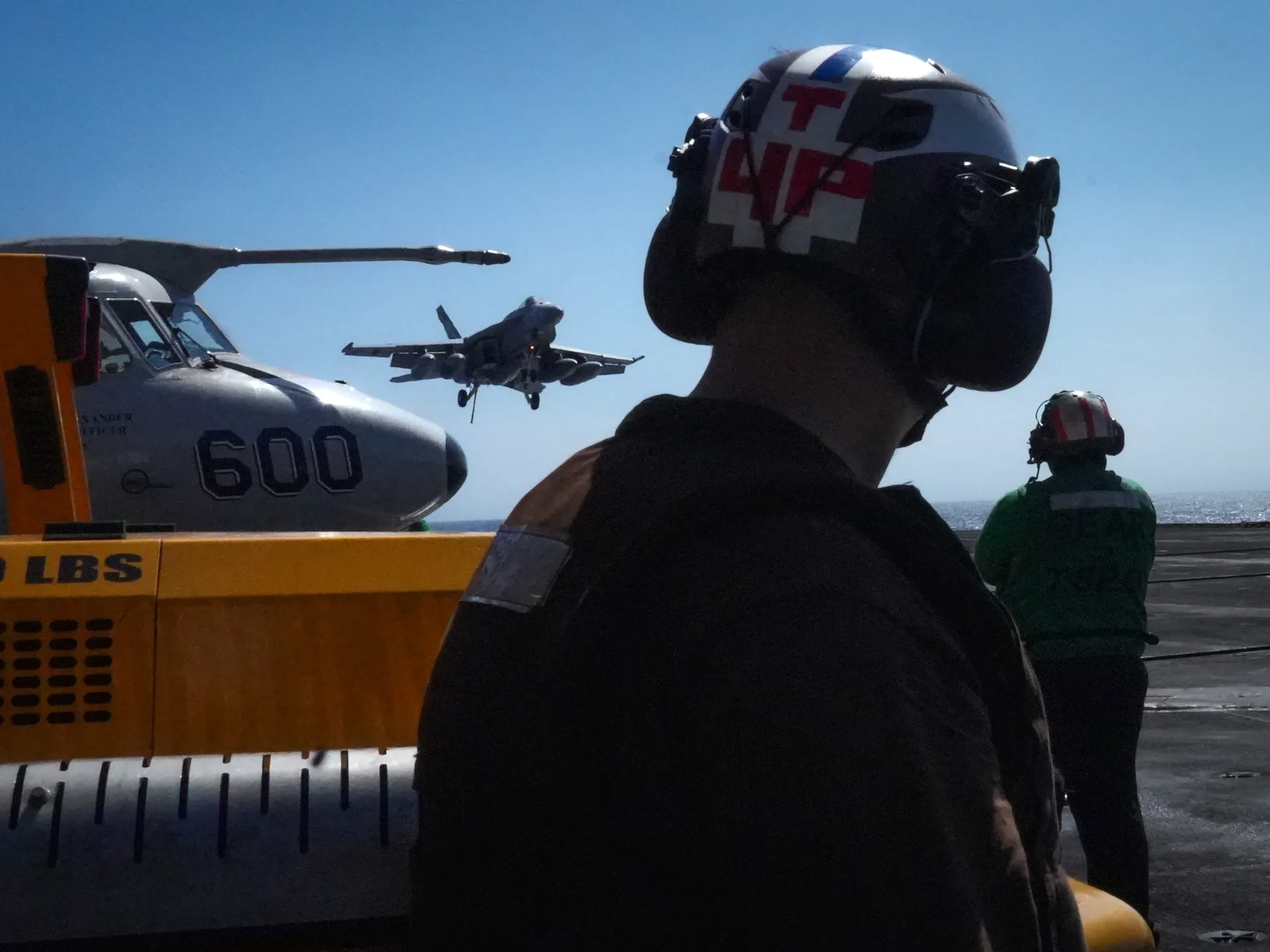 A flight deck crew member watches a military jet approach beside a parked aircraft and deck equipment.