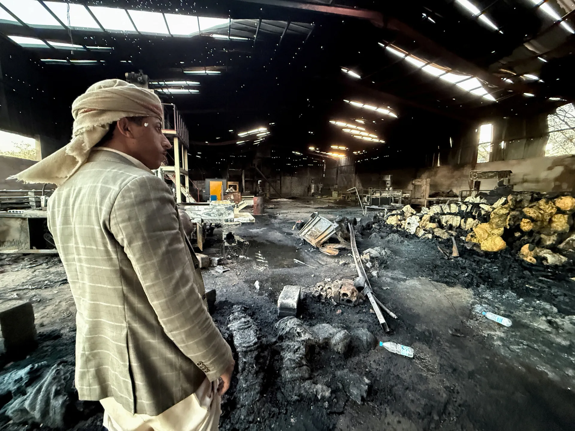 A man in a headscarf stands inside a fire-damaged workshop strewn with debris and burned materials.