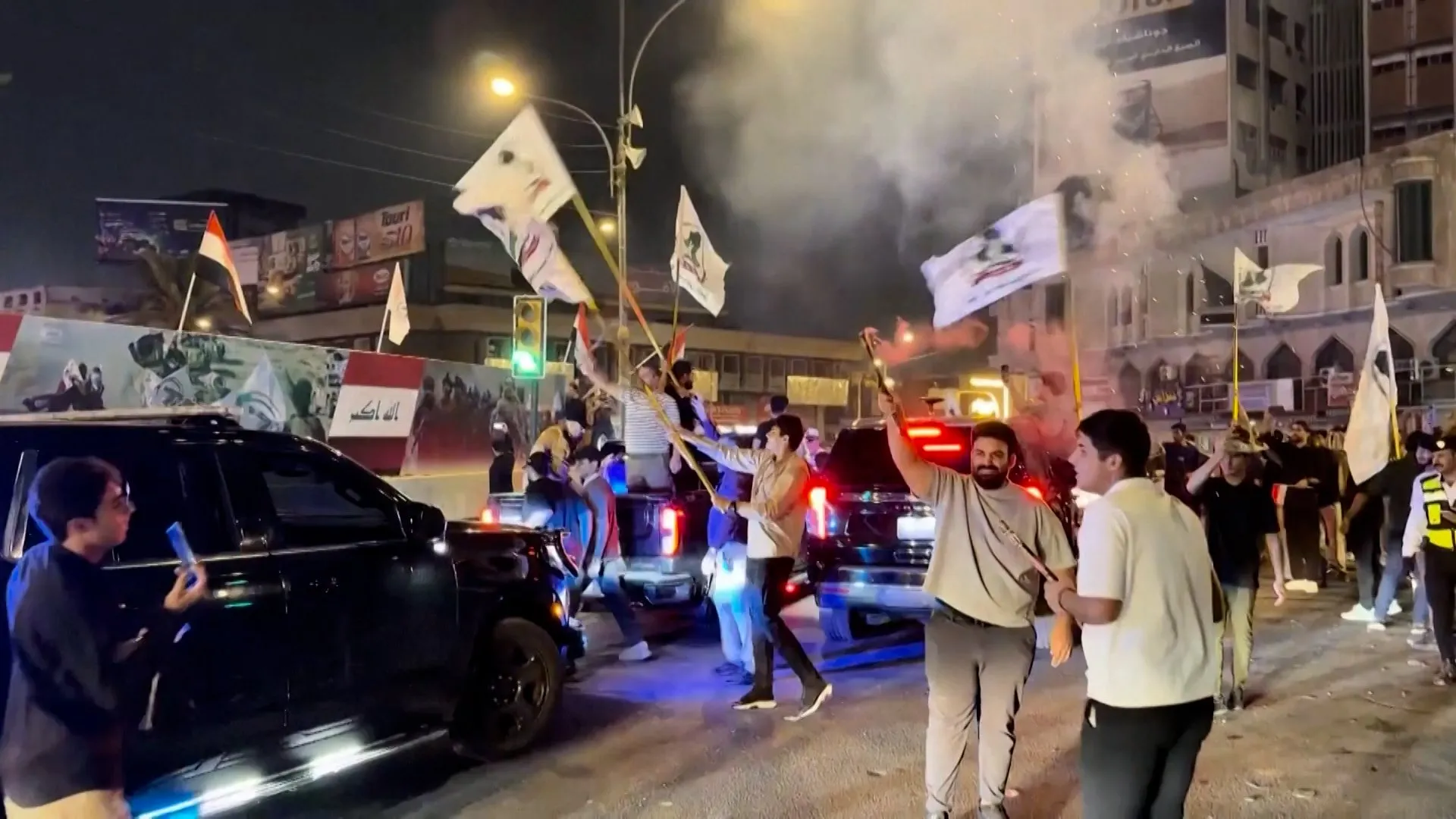 People wave flags beside vehicles on a city street at night as smoke rises nearby.