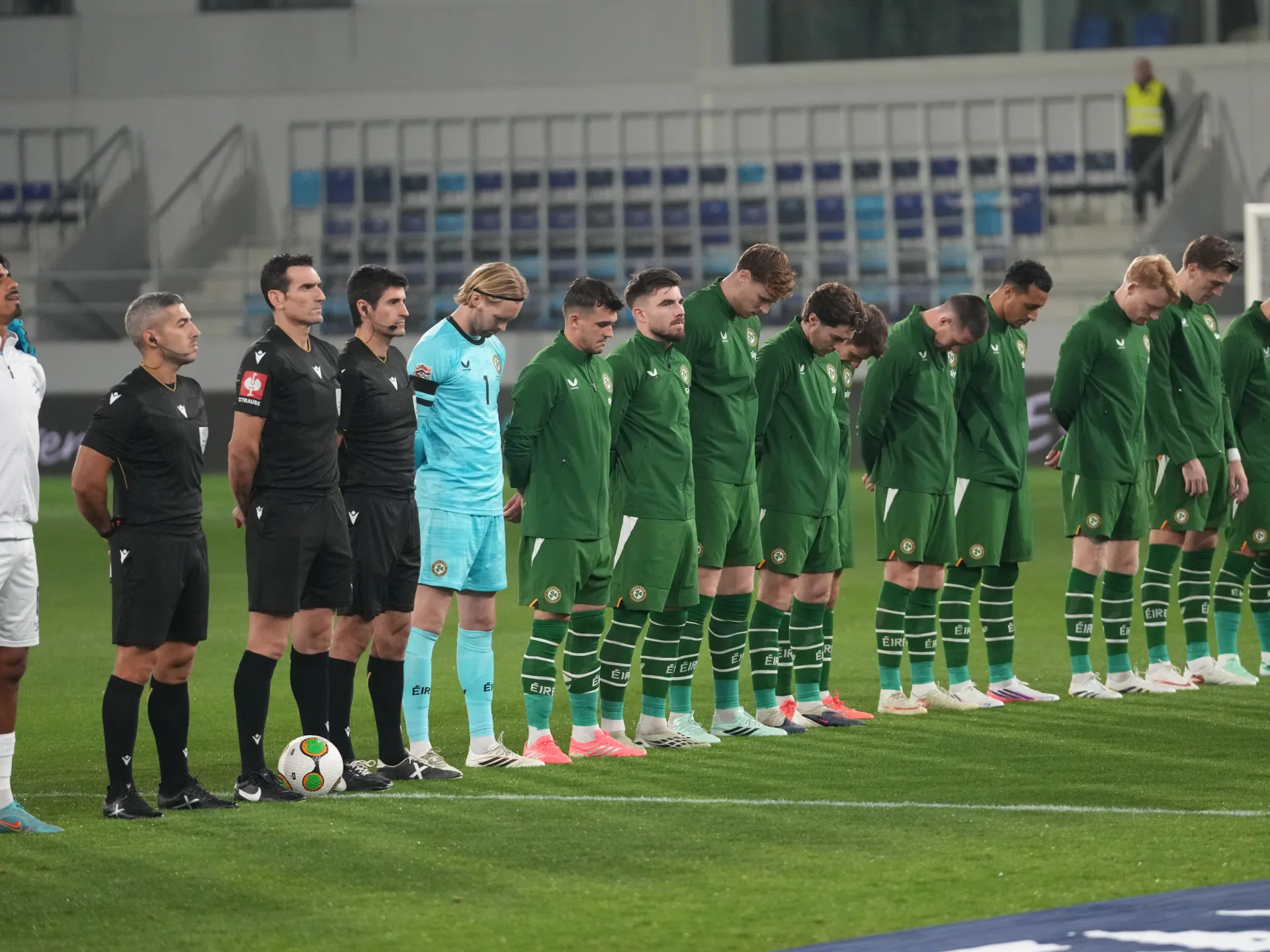 Irish players in green kits stand with heads bowed beside officials before a football match.