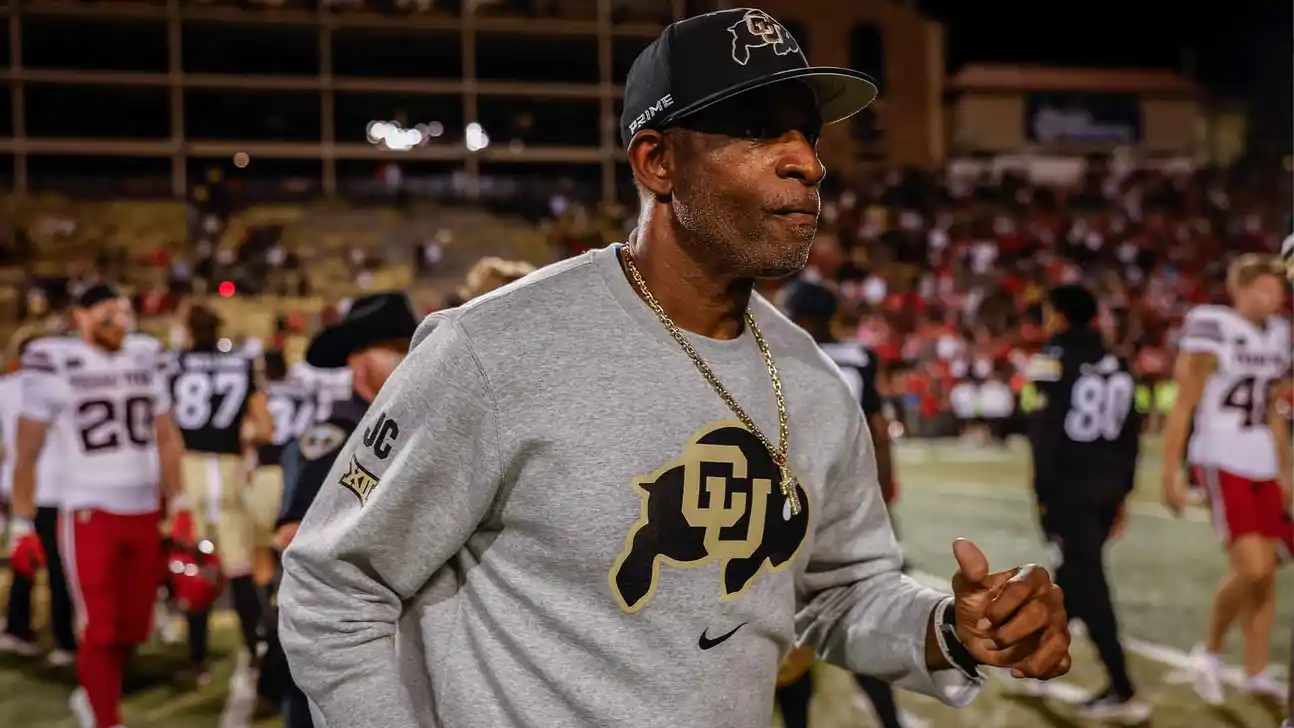 Man in a Colorado sweatshirt and black cap gestures on a football field during a game.