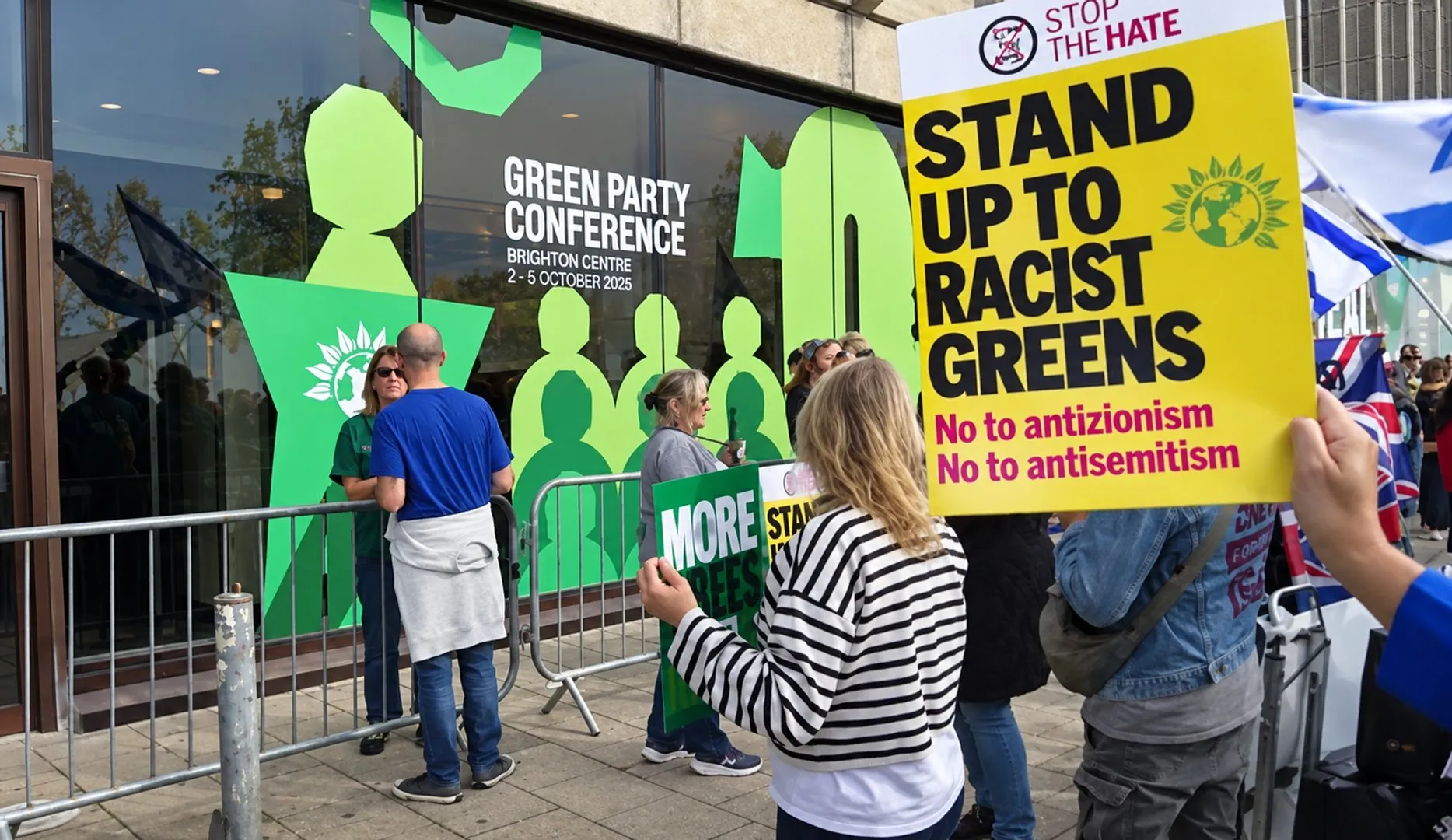 Protesters outside a Green Party conference hold signs opposing racism and antisemitism, with Israeli and Union Jack flags nearby.