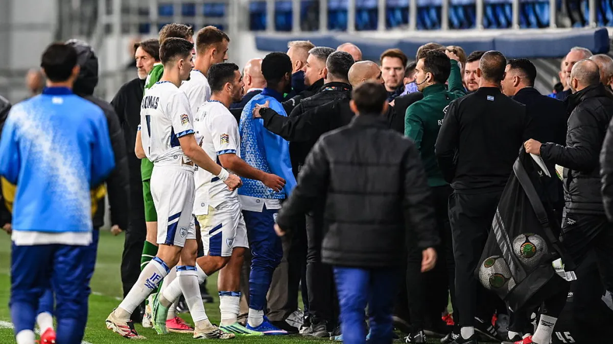 Football players and staff gather near the touchline, with a player in blue speaking to players in white.